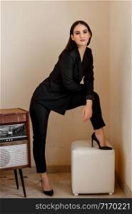 a beautiful Caucasian young woman in a black pantsuit and black sandals stands next to a vintage record player
