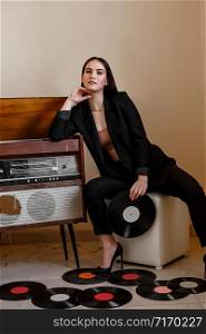 a beautiful Caucasian young woman in a black pantsuit and black sandals poses next to a vintage record player with a gramophone record in her hands