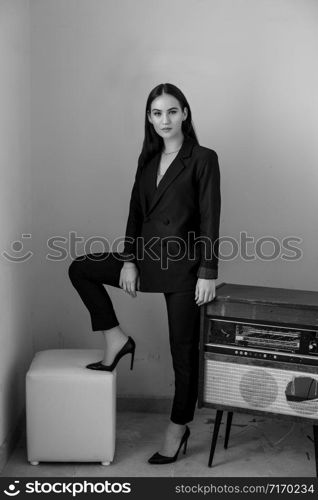 a beautiful Caucasian young woman in a black pantsuit and black sandals stands next to a vintage record player. black and white photo