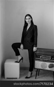 a beautiful Caucasian young woman in a black pantsuit and black sandals stands next to a vintage record player. black and white photo