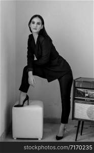 a beautiful Caucasian young woman in a black pantsuit and black sandals stands next to a vintage record player. black and white photo