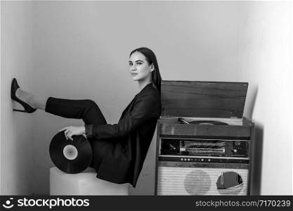 a beautiful Caucasian young woman in a black pantsuit and black sandals poses next to a vintage record player with a gramophone record in her hands. black and white image