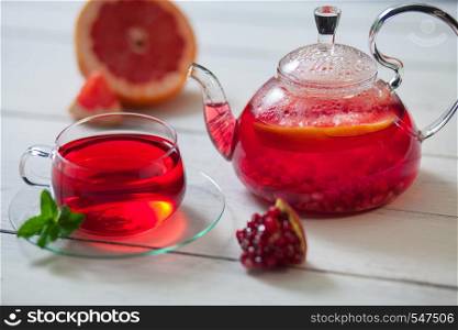 A beautiful composition of black tea and the addition of citrus. Glass teapot and cup of red tea with grape, pomegranate, mint on a white wooden table