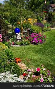 A beautiful, curving flower bed full of blooming annuals & perennials line a sunny backyard.