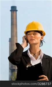 A beautiful dark haired woman wearing a hard hat and talking on her mobile phone while standing in front of a factory and its chimneys