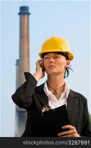 A beautiful dark haired woman wearing a hard hat and talking on her mobile phone while standing in front of a factory and its chimneys