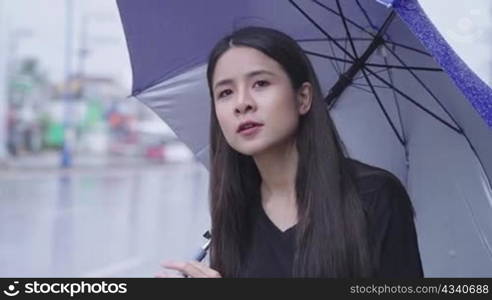 A beautiful east asian woman stands alone on roadside waiting for taxi or bus in hurry raining time, holding umbrella in rain, hurry rush hour, tropical climate change, urban style in rainy season