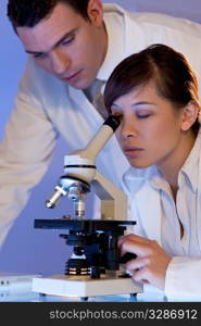 A beautiful female medical or scientific researcher using her microscope with her male colleague out of focus behind her.