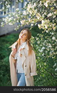 A beautiful girl in a long beige raincoat basks in the sun.. Portrait of a red-haired beauty on the background of a cherry blossom 296