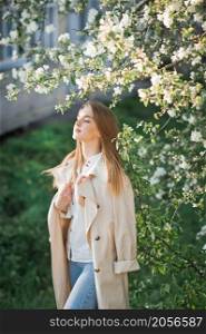 A beautiful girl in a long beige raincoat basks in the sun.. Portrait of a red-haired beauty on the background of a cherry blossom 296