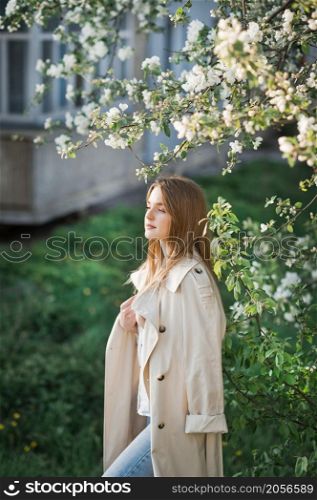 A beautiful girl in a long beige raincoat basks in the sun.. Portrait of a red-haired beauty on the background of a cherry blossom 296