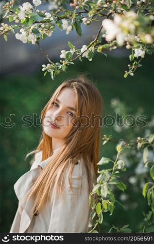 A beautiful girl in a long beige raincoat basks in the sun.. Portrait of a red-haired beauty on the background of a cherry blossom 297