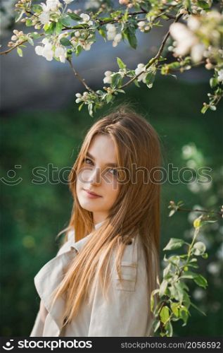 A beautiful girl in a long beige raincoat basks in the sun.. Portrait of a red-haired beauty on the background of a cherry blossom 297