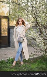 A beautiful girl in a long beige raincoat near a blooming cherry bush.. Portrait of a red-haired beauty in the spring nature 2946.