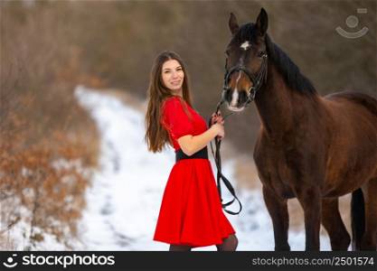 A beautiful girl in a red dress stands with a horse on the background of a winter road