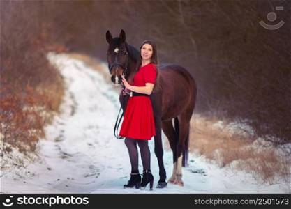 A beautiful girl in a red dress walks with a horse against the backdrop of a winter forest