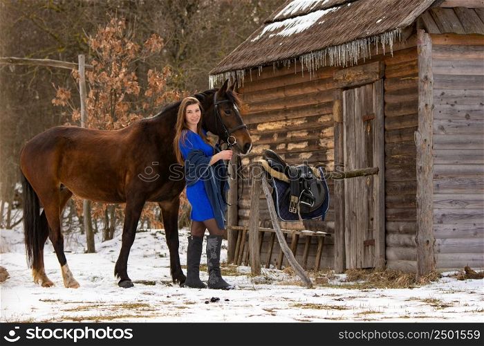 A beautiful girl in a short blue dress stands with a horse from an old wooden house