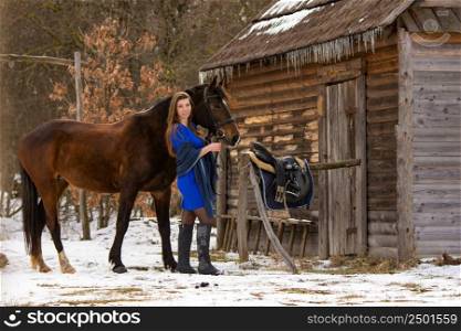 A beautiful girl in a short blue dress stands with a horse from an old wooden house