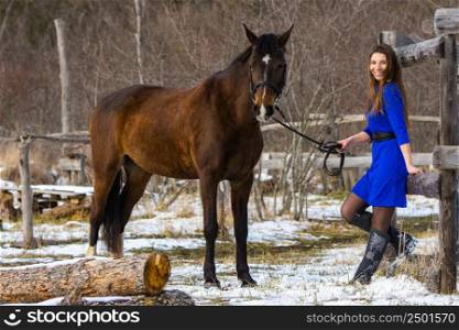 A beautiful girl in a short blue dress walks with a horse at an old farm in winter, the girl smiles and looks into the frame
