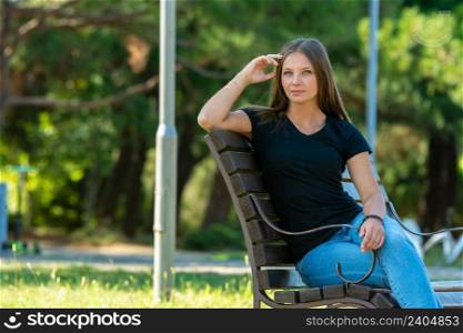 A beautiful girl in casual clothes sits on a bench in a beautiful green park, the girl looks into the frame