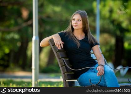 A beautiful girl in casual clothes sits on a bench in a beautiful green park