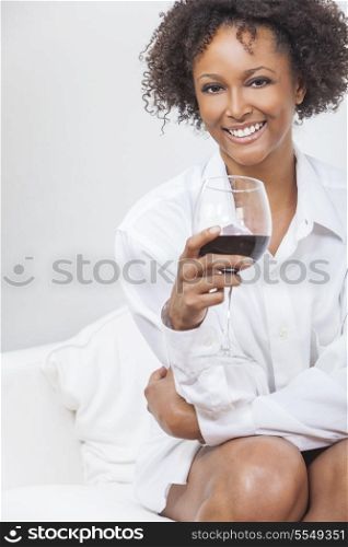 A beautiful happy mixed race African American girl or young woman wearing a white shirt and drinking red wine at home on a sofa