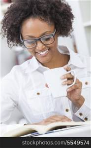 A beautiful happy mixed race African American girl or young woman wearing glasses drinking coffee or tea and reading a book in her kitchen at home