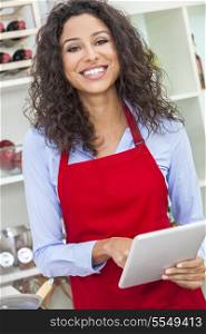 A beautiful happy young woman or girl wearing a red apron &amp; using a tablet computer while cooking in her kitchen at home