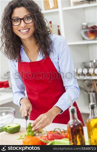 A beautiful happy young woman or girl wearing glasses &amp; a red apron cutting &amp; preparing fresh vegetable salad food in her kitchen at home