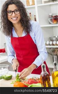 A beautiful happy young woman or girl wearing glasses &amp; a red apron cutting &amp; preparing fresh vegetable salad food in her kitchen at home