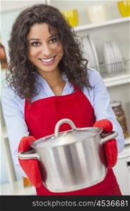 A beautiful Latina girl or young woman looking happy wearing red apron, cooking holding metal pot in her kitchen at home