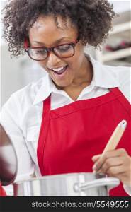 A beautiful mixed race African American girl or young woman looking happy wearing glasses a red apron &amp; cooking in her kitchen at home