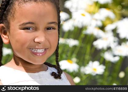 A beautiful mixed race little girl smiles while outside in glorious summer sunshine and backed by giant daisies
