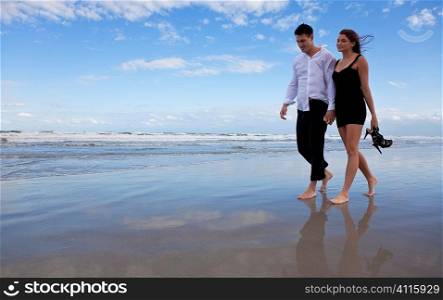A beautiful romantic young man and woman couple walking hand in hand and barefoot on a beach. They are dressed as if it is after a night out and the girl is carrying her shoes.