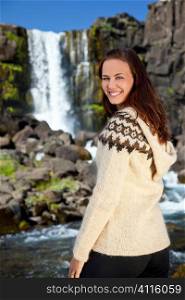 A beautiful Scandinavian woman wearing traditionally patterned knitwear smiling in front of a mountain waterfall. Shot on location in Iceland.