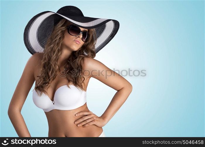 A beautiful sexy girl posing in white bikini and stylish sun hat on blue studio background.