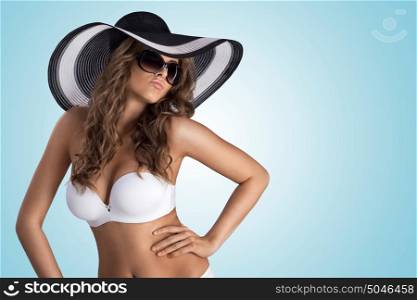 A beautiful sexy girl posing in white bikini and stylish sun hat on blue studio background.