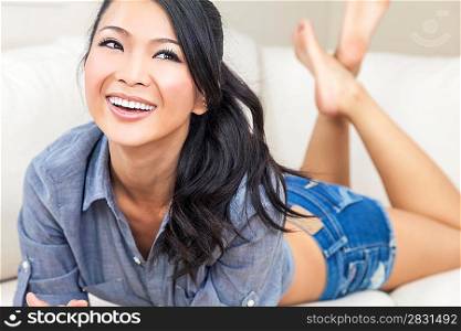 A beautiful sexy young Chinese Asian Oriental woman with a wonderful toothy smile relaxing on a sofa in denim jeans shorts