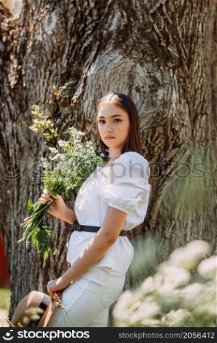A beautiful slender girl with a bouquet of wildflowers on the background of a summer park.. A walk of a joyful girl with a bouquet of daisies in a summer park 3564.