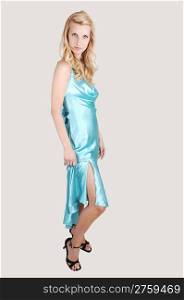 A beautiful tall blond woman in an light blue long dress and high heelsstanding from the site in the studio for a portrait, on light gray background.