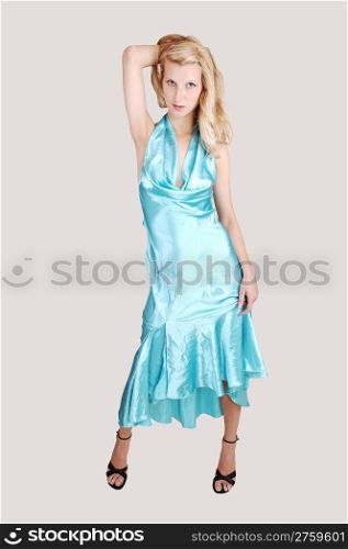 A beautiful tall blond woman in an light blue long dress and high heelsstanding from the front in the studio for a portrait, on light gray background.