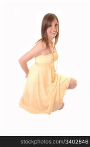 A beautiful teenager in a yellow dress kneeling on the floor in the studioholding her hands behind her back and smiling into the camera, over white.