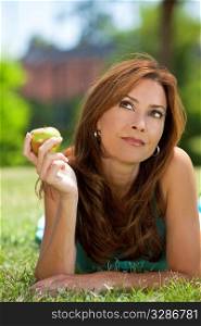 A beautiful woman in her thirties laying down outside on grass holding or eating an apple looking up and thinking