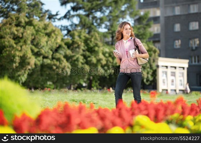 A beautiful woman with a mobile phone and coffee to go walking in a city park.