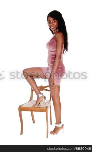 A beautiful young African woman standing in a short pink dress andhigh heels, one leg up on a chair, isolated for white background