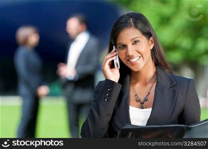 A beautiful young Asian businesswoman with a wonderful smile chatting on her cell phone with her colleagues out of focus behind her.