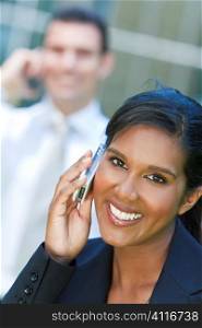 A beautiful young Asian businesswoman with a wonderful smile chatting on her cell phone with an out of focus male colleague behind her.