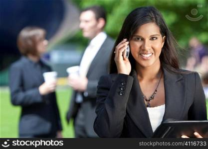 A beautiful young Asian businesswoman with a wonderful smile shot using her cell phone outside with her colleagues out of focus behind her.