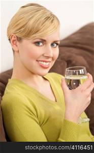 A beautiful young ble eyed blond woman sitting on a settee drinking a glass of water.