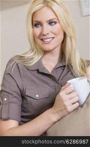 A beautiful young blond woman drinking tea or coffee from a white mug sitting at home on a her sofa